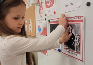 Dziewczynka przyczepia na tablicy ilustrację Marii Skłodowskiej - Curie