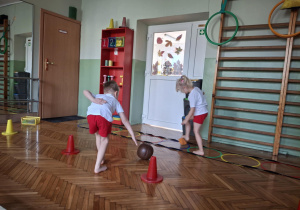 Chłopiec turla piłkę a dziewczynka podnosi woreczek gimnastyczny