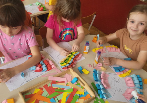 dziewczynki siedzą przy stole i składają i przyklejaja kolorowe paski