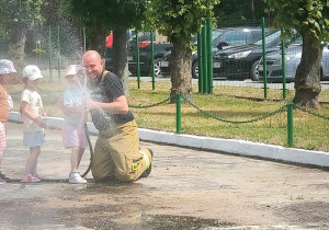 dziewczynka ze sgtrażakiem leje wodę z węża strażackiego