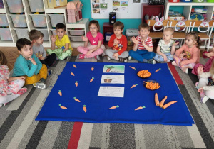 dzieci z grupy pierwszej i drugiej siedzą na dywanie przed rozłożonymi planszami z ilustracjami marchewki i jej uprawą