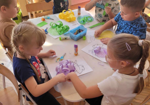 Dzieci siedzą przy stoliku i kolorują szablon owocu i wycinają paski z kolorowego papieru