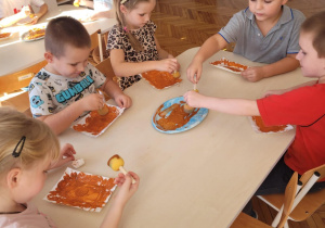 Dzieci siedzą przy stoliku i malują brązową farbą talerzyk papierowy