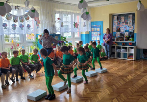 dzieci z grupy trzecej ubrane w zielone stroje do tańca, prezentują układ taneczny na steach przed siedzącymi dziećmi z przedszkola miejskiego nr 114