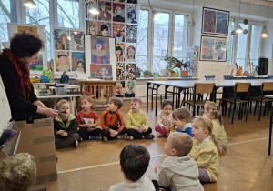 Dzieci siedzą w pracowni plastycznej i słuchaja pani plastyk