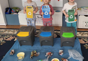 trzehc chłopców stoi i pokazuje kolory pojemników do segregacji odpadów - papier niebieski, plastik żółty i szkło zielony