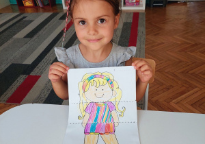 dziewczynka siedzi przy stoliku i prezentuje pokolorowany rysunek postaci dziecka
