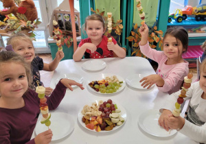 pięć dziewczynek siedzi przy stoliku i pokazuje zrobioneszaszłyki z owoców