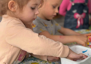 dwoje dzieci macza palce w farbbie, a trzecie dziecko na nie patrzy