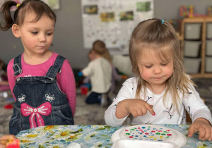 dziewczynka maluuje palcem zamoczonym w farbie swój talerzyk, druga dziewczynka stojąc na nią patrzy