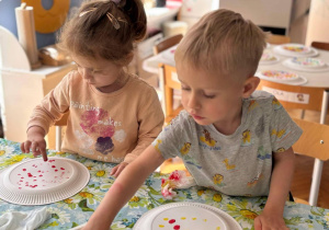 dwoje dzieci siedząc przy stoliku malują kropki na tlerzykavch farbami