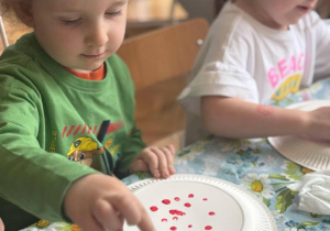dwoje dzieci siedząc przy stoliku malują kropki na tlerzykavch farbami