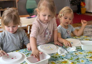 troje dzieci siedząc przy stoliku malują kropki na tlerzykavch farbami