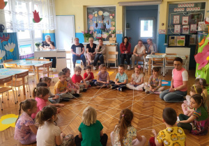 na zdjęciu widać siedzące w kole na podłodze dzieci, które rozwijają pajęczynę z włóczki podczas zabawy wyciszającej oraz p.dyrektor i radę pedagogiczną, które oglądają zajęcie