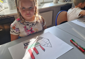 Dziewczynka siedzi przy stole z pokolorowanym rysunkiem statku
