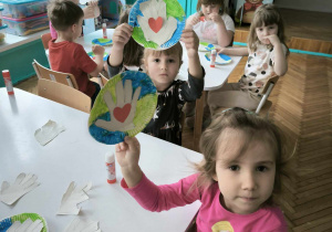 dwójka dzieci siedzi przy stoliku i prezentuje swoją pracę plastyczną wykonaną z talerzyka papierowego ma którym jest namalowana planeta ziemia