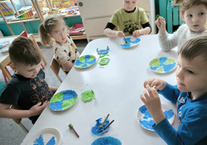 dzieci siedząprzy stolikach i malują talerzyki papierowe na zielono i niebiesko tworząc planetę
