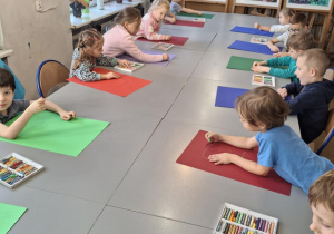 Dzieci siedzą przy stolikach z kolorowymi dużymi kartonami