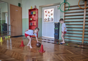 Dziewczynka toczy piłke a chłopiec podnosi stopą woreczek