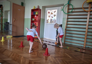 Chłopiec turla piłkę a dziewczynka podnosi woreczek gimnastyczny