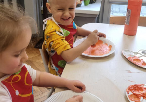 dzieci siedząc przy stoliku maluja pomarańczowa farbę talerzyki papierrowe