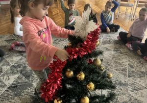 Dziewczynka zakłada czerwony łańcuch choinkowy na choinke