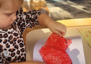 dziecko wykonuje pracę plastyczną, odkleja serce z folii bąbelkowej pozostawiając wzór serca na kartonie