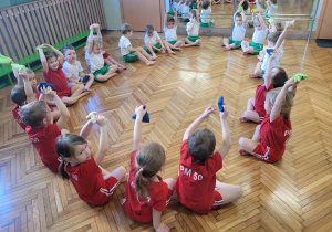 Dzieci siedzą w siadzie skrzyżnym i podnoszą woreczek do góry