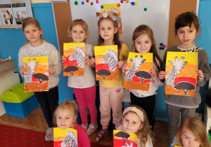 dziewczynki prezentują pracę plastyczną "Na sawannie" wykonana farbami i naklejoną sylwetą zebry oraz żyrafy w kolorach czarnobialych